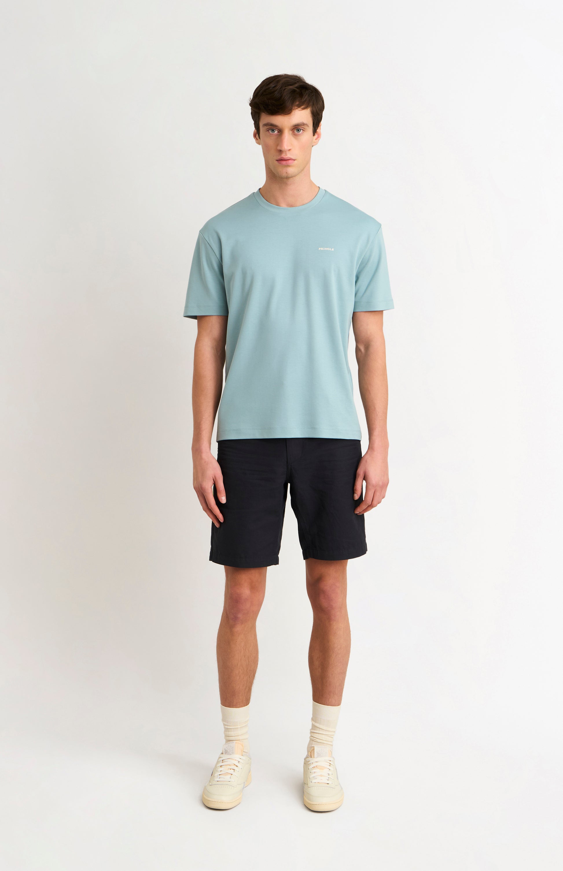 Man wearing a blue t-shirt and black shorts and white trainers on a white background