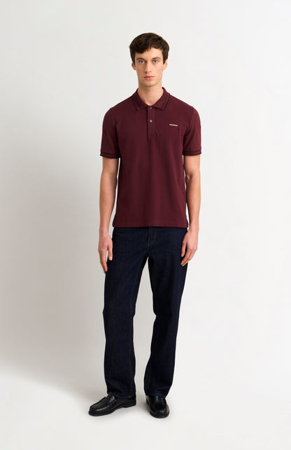 Model wearing a burgundy polo shirt with black tipping details and dark jeans against a light grey background