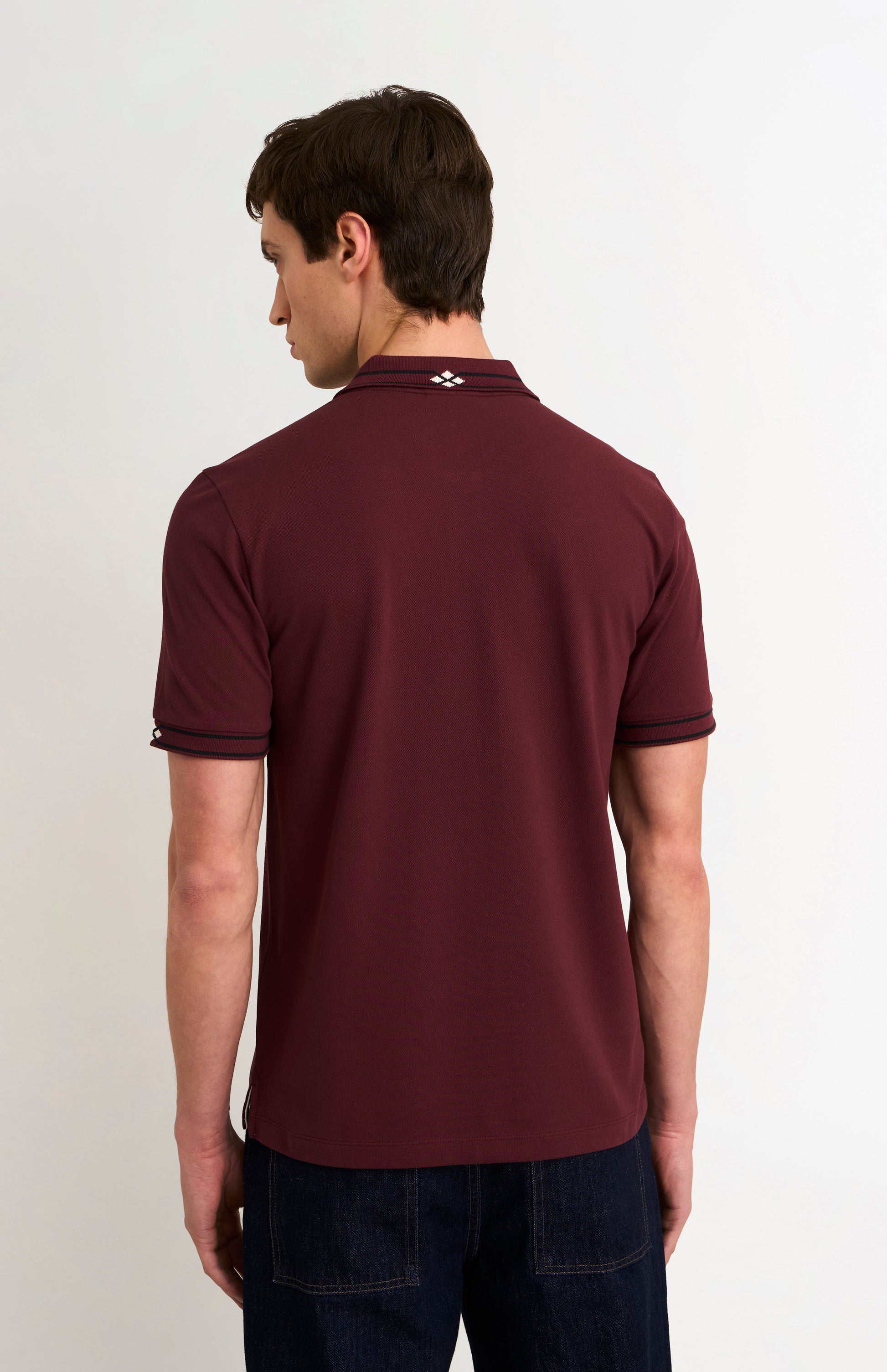Back profile of model wearing a burgundy polo shirt with black tipping details and dark jeans against a light grey background