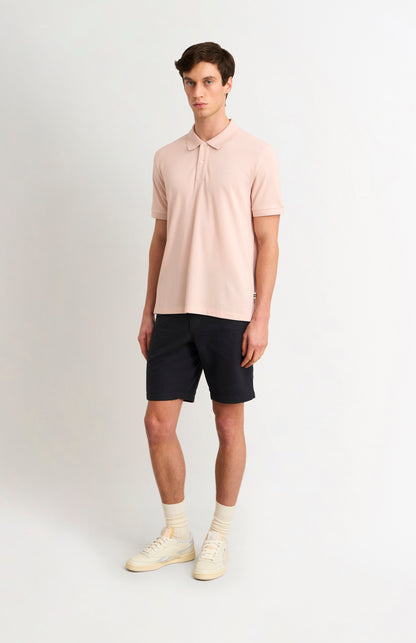 Man wearing dark shorts and a dusty pink polo shirt with white trainers on a white background