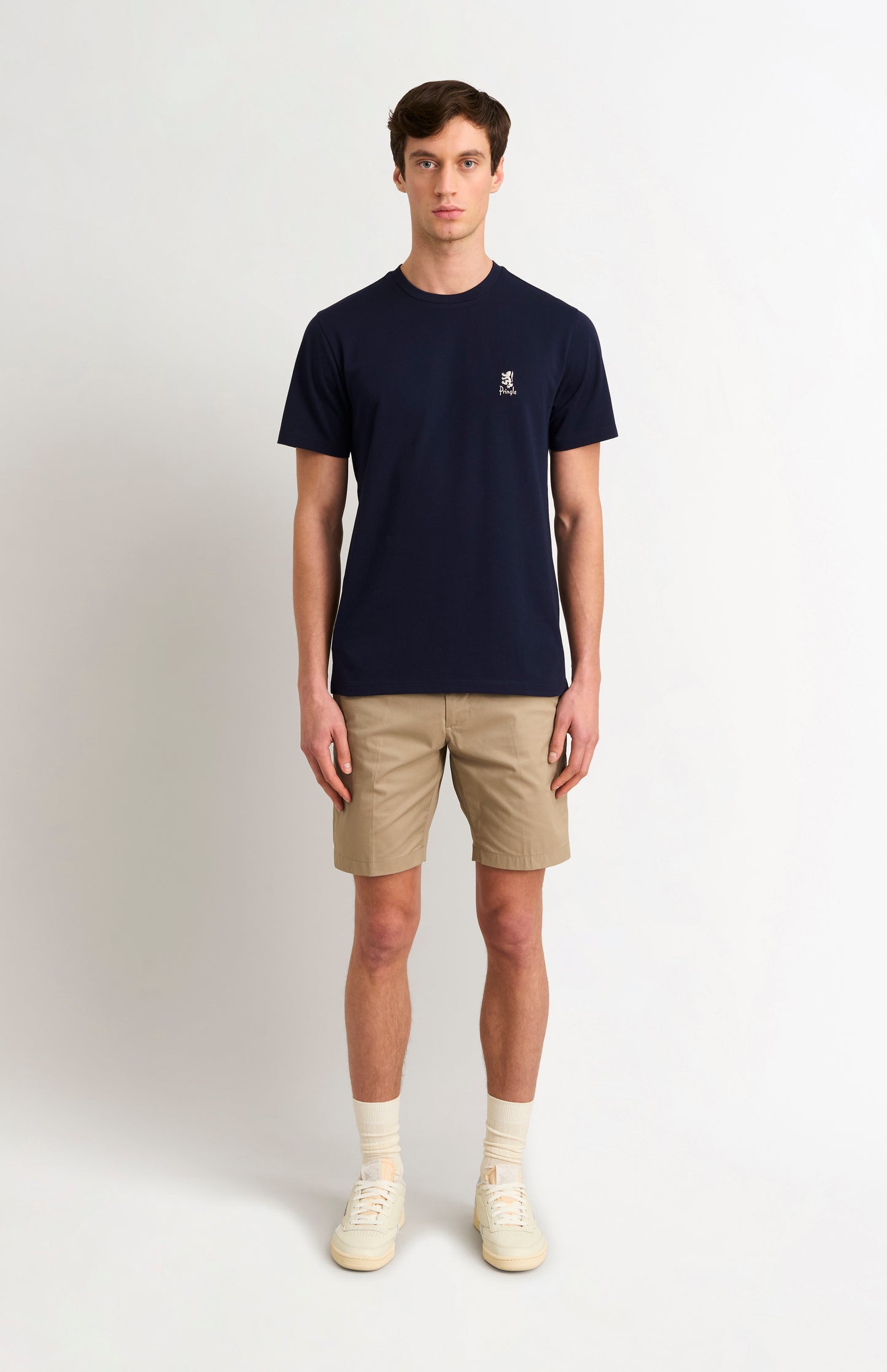 Man wearing a navy t-shirt with a small logo and beige shorts on a white background