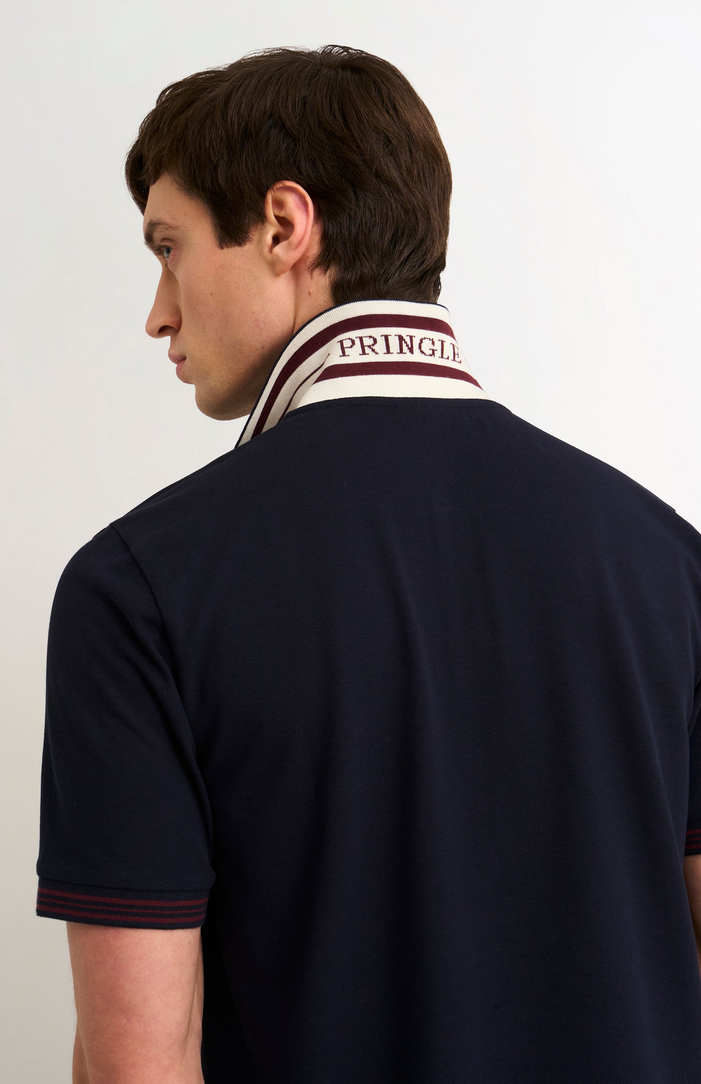 Detail shot of model wearing a navy polo shirt with burgundy tipping details and Pringle written under flipped-up collar