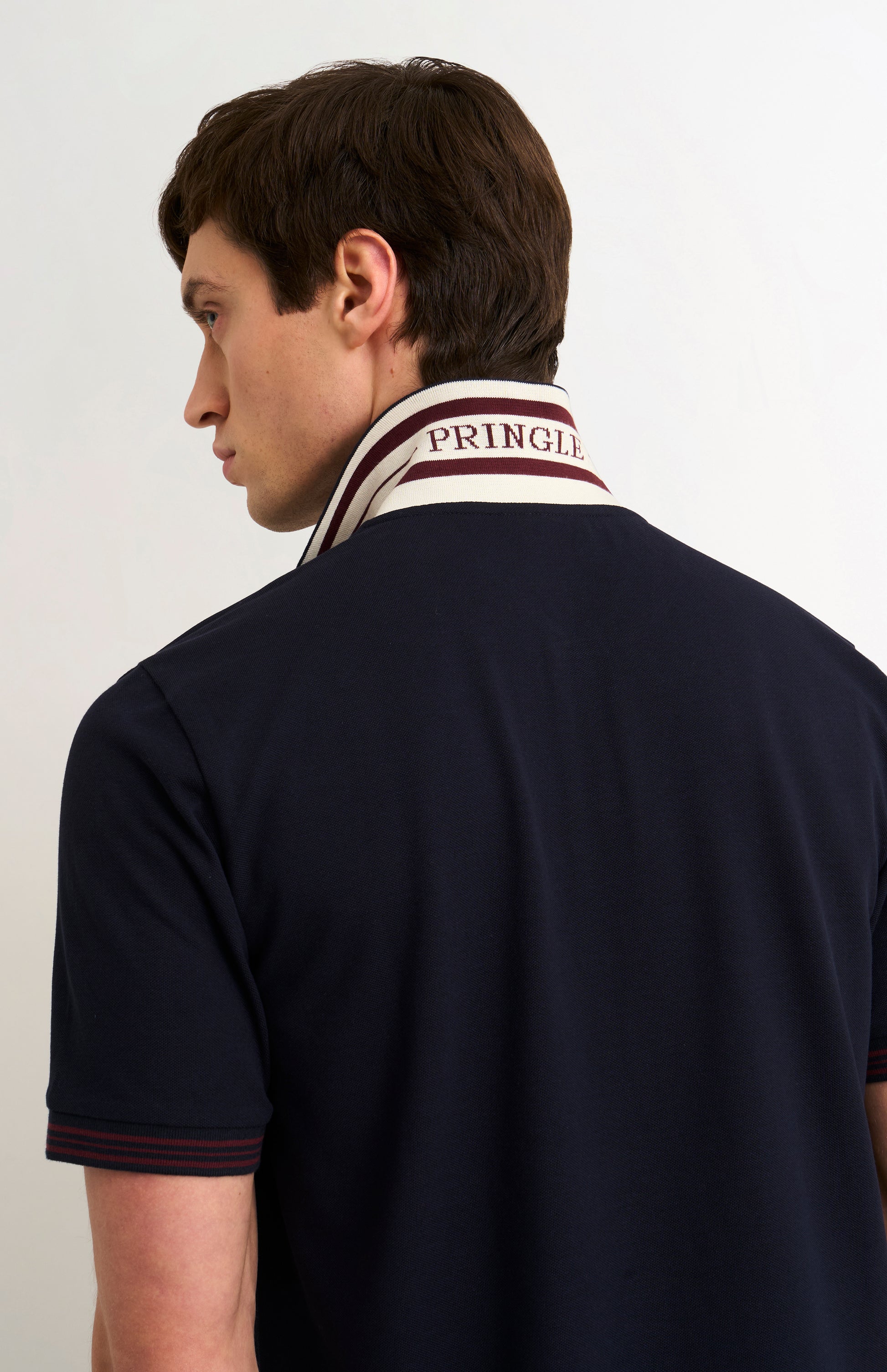 Detail shot of model wearing a navy polo shirt with burgundy tipping details and Pringle written under flipped-up collar
