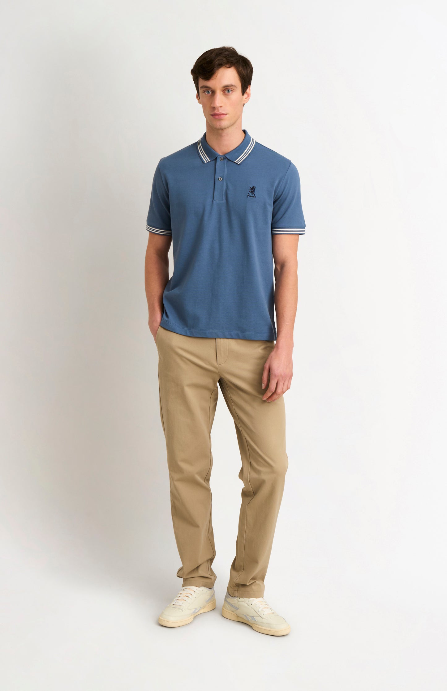 Full body shot of model wearing a mid blue polo with tipping details and Pringle logo on upper side of chest