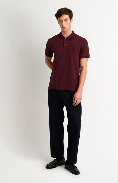 Model wearing a burgundy polo with black tipping and a small Pringle logo on top side of chest, and dark trousers against a light grey background