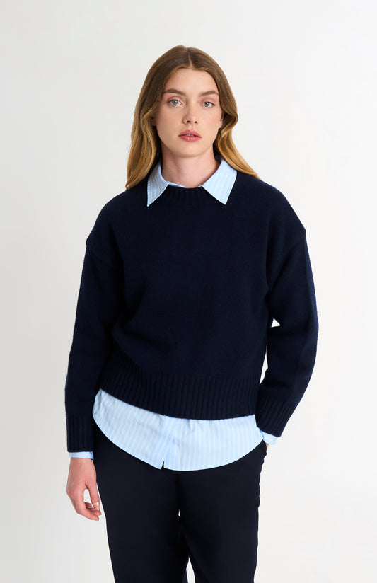 Woman wearing a navy sweater over a white shirt with a plain background