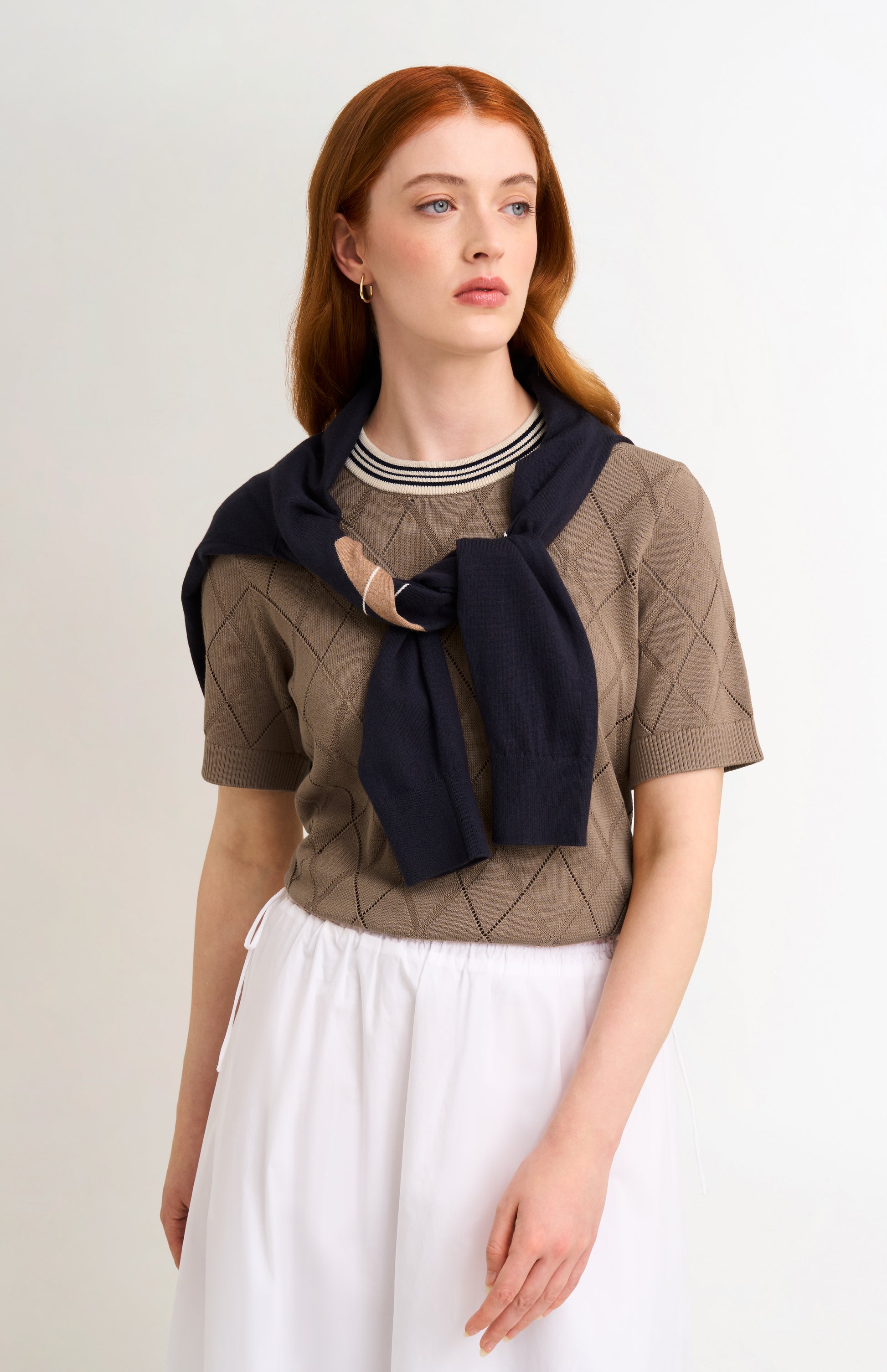 A model with red hair wearing a brown argyle knit short-sleeve top with a navy blue scarf tied around the neck, paired with a white skirt, against a plain light background.
